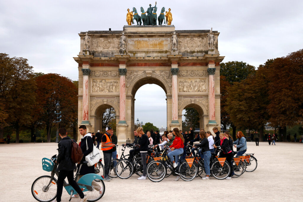 A group of tourists on bicycles take a cycling break in front of the Arc de Triomphe du Carrousel next to the Louvre museum in Paris, France